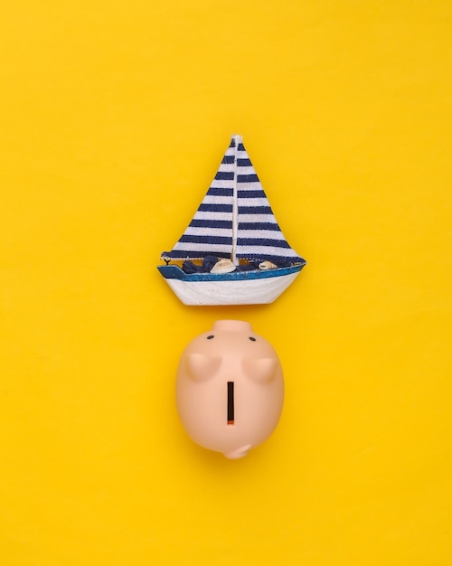 Sailboat and piggy bank on yellow background. Travel concept. Top view