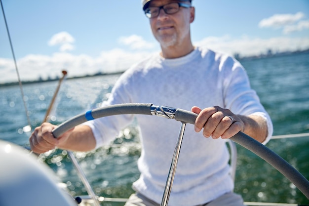 sailing, age, tourism, travel and people concept - close up of happy senior man in captain hat steering wheel and navigating sail boat or yacht floating in sea