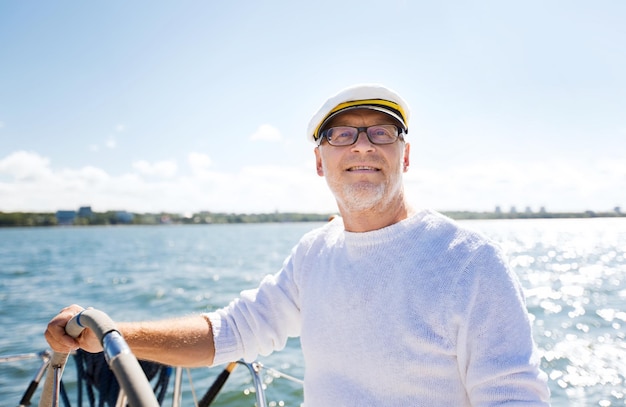 sailing, age, tourism, travel and people concept - happy senior man in captain hat on steering wheel and navigating sail boat or yacht floating in sea