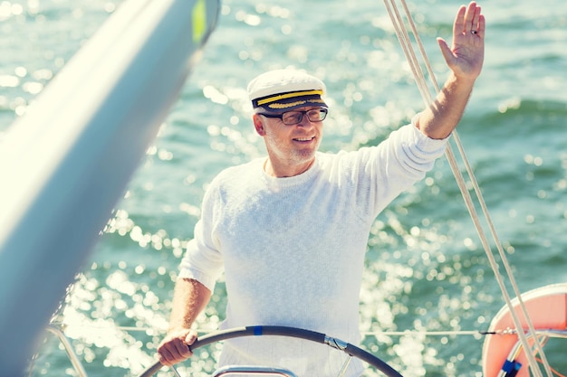 Photo sailing, age, tourism, travel and people concept - happy senior man in captain hat on steering wheel and navigating sail boat or yacht floating in sea