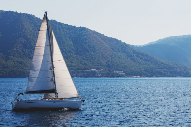 Sailing yacht with white sails on a wavy sea bay on a background of mountains