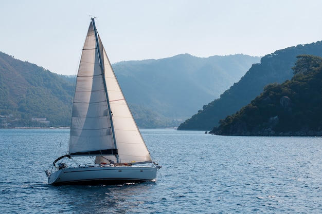 Sailing yacht with white sails on a wavy sea bay on a background of mountains