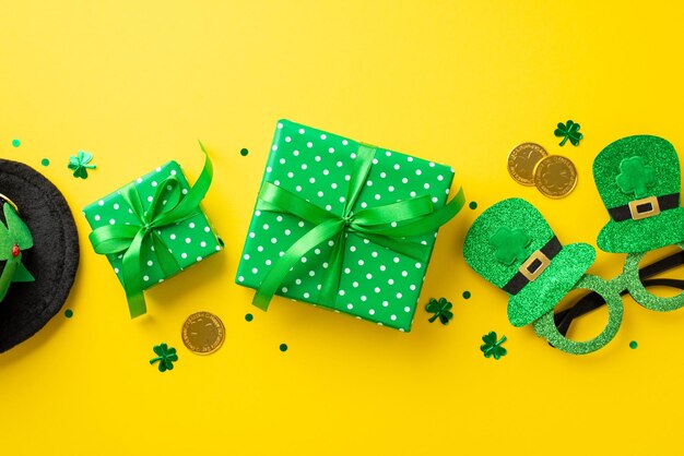 Saint Patricks Day concept Top view photo of green gift boxes with ribbon bows gold coins hat shaped party glasses leprechaun headwear and trefoil shaped confetti on isolated yellow background