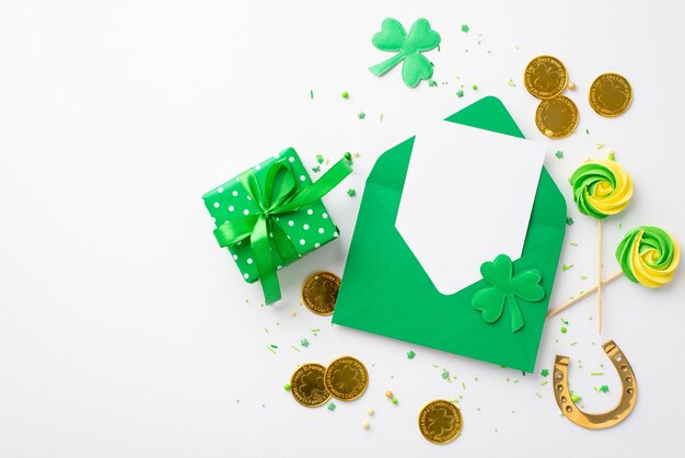 saint patricks day concept top view photo of open green envelope with paper sheet meringue lollipops giftbox gold coins horseshoe shamrocks and sprinkles on isolated white background with copyspace
