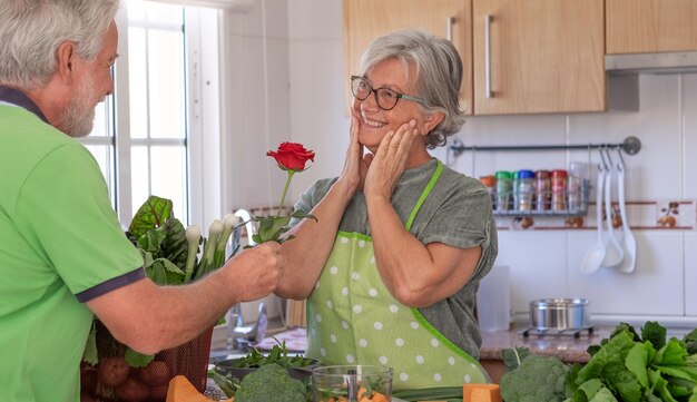 聖バレンタインの日 年配の夫が妻に赤いバラを提供している 家庭の台所で野菜を準備している美しい老夫婦