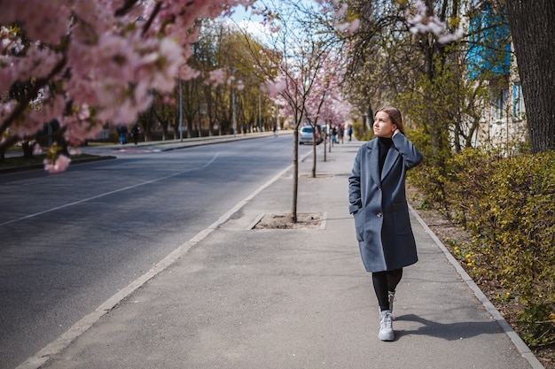 Ветки сакуры с цветами на дереве на улицах города. Счастливая женщина-девушка в серой палитре идет по аллее с цветущей сакурой. Великолепная модная девушка на открытом воздухе. Цветущее дерево сакуры.