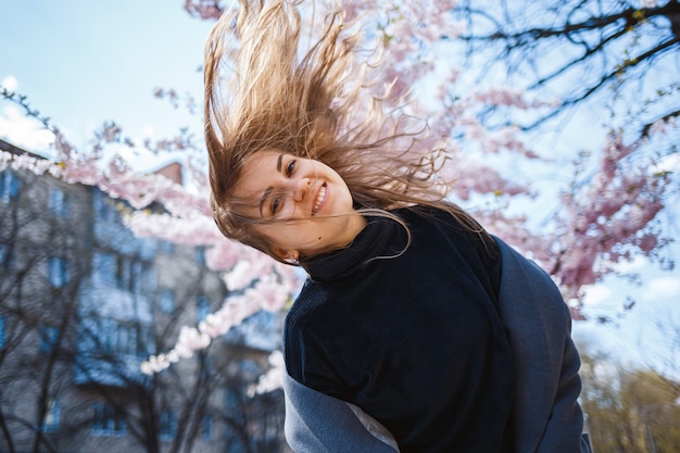 街の通りの木に花が咲く桜の枝。桜が咲く通りで回転する幸せな女の女の子。屋外でゴージャスなファッショナブルな女の子。桜。