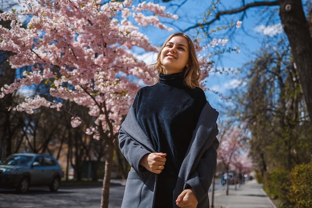 Sakura branches with flowers on a tree on the city streets. Happy woman girl spinning on the street with blooming sakura. Gorgeous fashionable girl outdoors. Sakura tree blossoms.