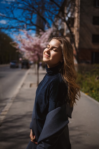 Sakura takken met bloemen aan een boom in de straten van de stad. Stijlvolle vrouw meisje loopt op straat met bloeiende sakura. Schitterend modieus meisje buitenshuis. Sakuraboom bloeit.