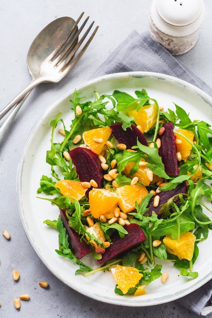 Insalata di rucola, barbabietola al forno, arancia e cipolle rosse con spezie e pinoli in un piatto bianco. olio d'oliva e ingredienti sulla tavola di pietra grigia. messa a fuoco selettiva.