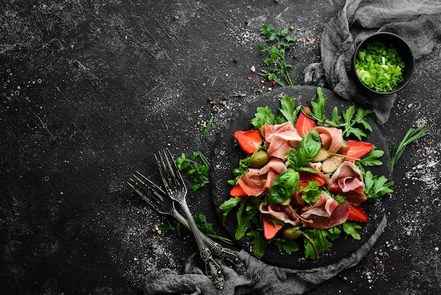 Салат из рукколы, прошутто, клубники и каперсов на черной каменной тарелке итальянской кухни