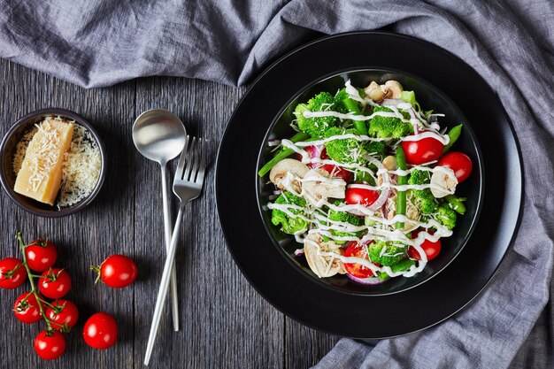 Salad of broccoli florets, green beans, champignons, tomatoes, red onions topped with grated parmesan cheese and yogurt dressing in a black bowl, landscape view from above, flat lay