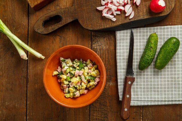 Salad in a clay plate on a table on a wooden table next to chopped vegetables and sausage on the planks