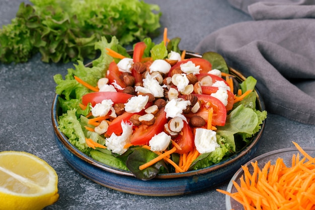 A salad of fresh lettuce leaves, tomato,mozzarella, carrots and hazelnut with olive oil and juice lemon in a ceramic plate on a gray background.