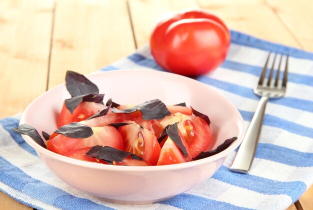 Salad of fresh tomatoes with basil leaves in a bowl on napkin on wooden background