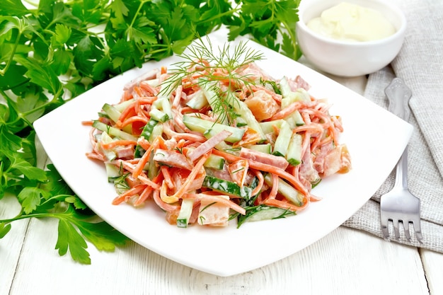 Insalata di salsiccia affumicata, carota piccante, pomodoro, cetriolo e spezie, condita con maionese, asciugamano, forchetta e prezzemolo su uno sfondo di tavola di legno chiaro