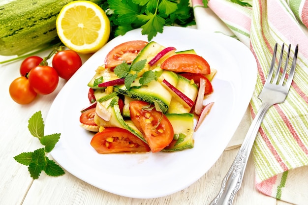 Salad from young zucchini radish tomato and mint flavored with lemon juice and soy sauce in a white plate napkin and fork on a wooden plank background