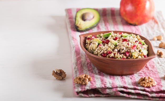 Salad of germinated buckwheat, avocado, walnut and pomegranate seeds in clay plate on white wooden background.