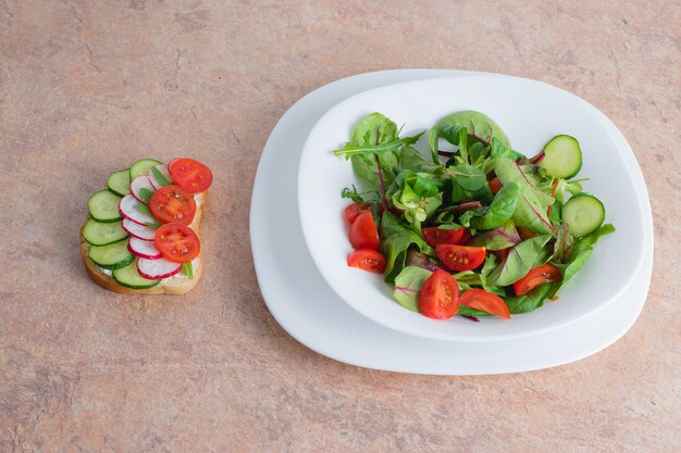 Insalata di verdure verdi e pomodoro in un piatto bianco sul tavolo. insalata di verdure primaverili con un panino di verdure su uno stle in un piatto bianco.