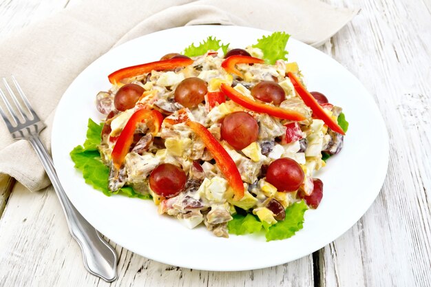 Salad of meat, salty soft feta cheese, sweet pepper, egg and grape with mayonnaise on lettuce the plate, napkin and fork on the background light wooden boards