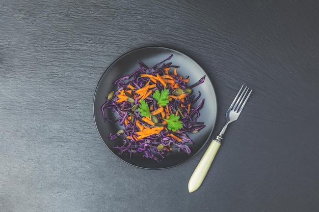 Фото Салат из фиолетовой капусты, моркови, тыквенных семечек и петрушки в черной керамической тарелке на поверхности черного камня, вид сверху, копия специй
