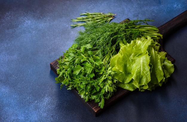 Salad parsley and dill on a dark cutting board against a blue concrete background