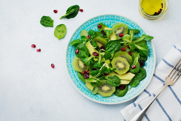 Salad of spinach baby leaves, watercress, kiwi, avocado and pomegranate in blue ceramic plate on white light wooden surface. Selective focus. Top view. Copy space.