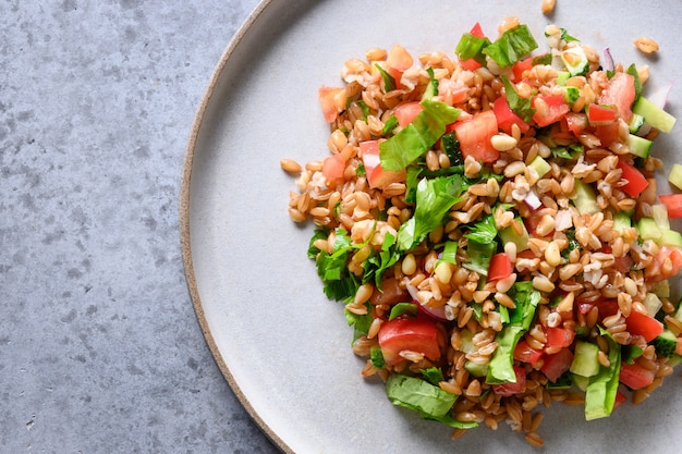 Salad of whole grain cereal spelt with vegetables, tomato, cucumber, greens on grey stone table with 