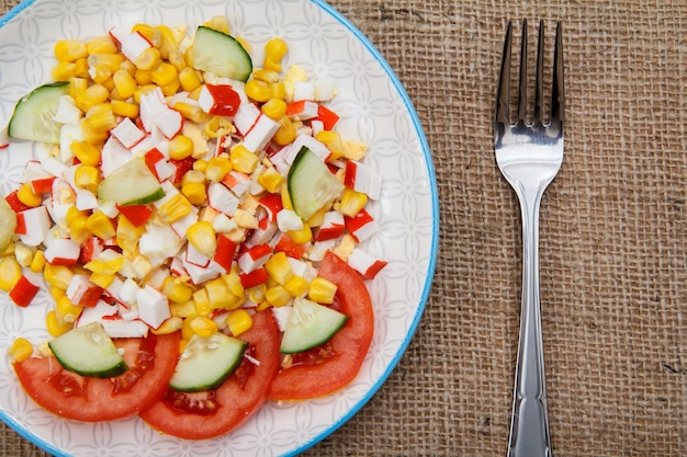 Салат с крабовыми палочками, вареной кукурузой и свежесрезанными помидорами и огурцами на тарелке с вилкой на мешковине. Вид сверху.