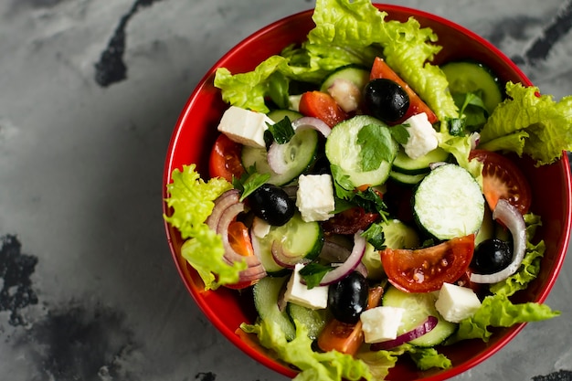Photo salad with fresh vegetables, cheese and olives in a red plate, on a gray background