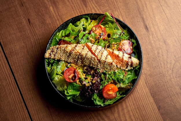 Salad with greens cherry tomatoes and grilled salmon in a black plate Wooden background