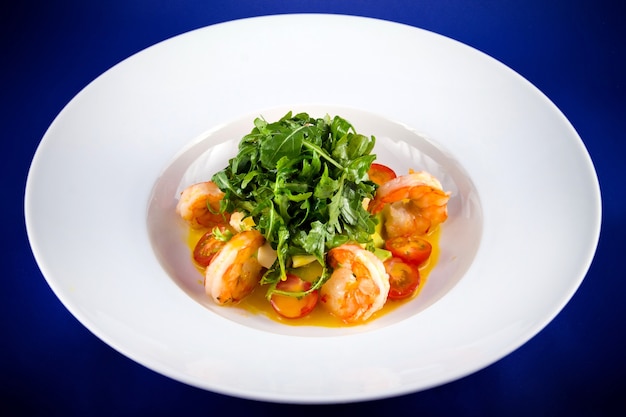 Salad with king prawns, arugula, tomatoes, avocado. Flat lay top view on a white plate on a blue background.