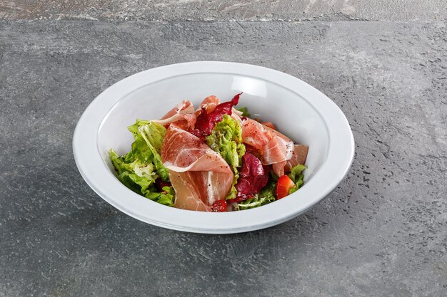 Salad with prosciutto in white plate on grey concrete background