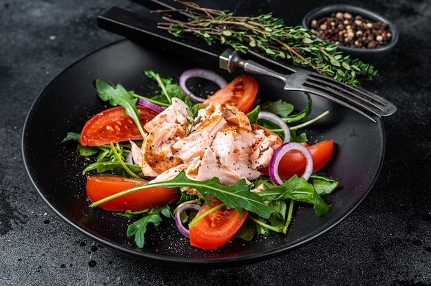 Salad with roasted salmon fillet steak, fresh salad arugula and tomato in a plate. Black background. Top view.