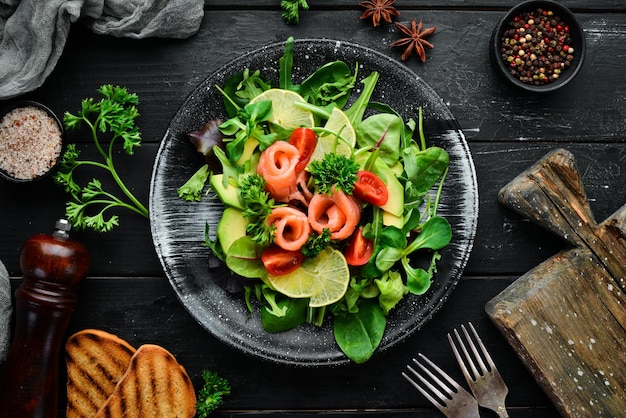 Insalata con salmone, avocado, pomodori e spinaci in un piatto nero su sfondo di legno vista dall'alto spazio libero per il testo disposizione piatta