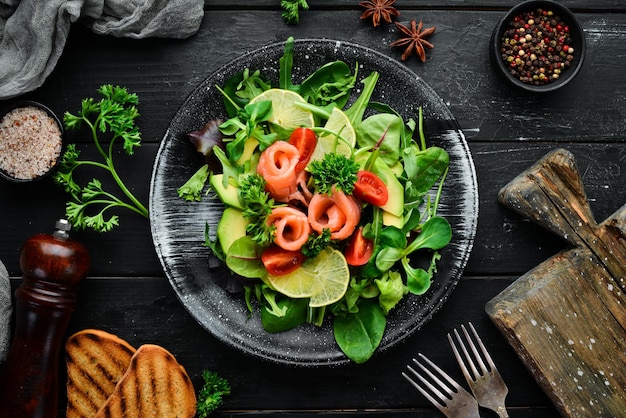 Salad with salmon avocados tomatoes and spinach In a black plate on a wooden background Top view Free space for your text Flat lay