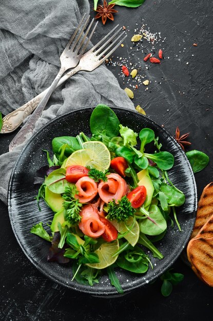 salad with salmon avocados tomatoes and spinach in a black plate on a wooden background top view free space for your text flat lay