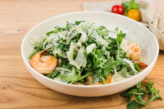 Salad with shrimp, arugula and parmesan cheese in a plate on a light wooden surface.