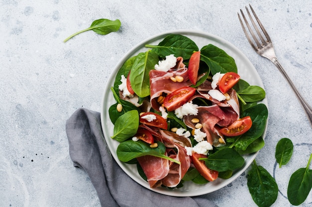 Salad with spinach, cherry tomato, mozzarella, pine nuts and ham with olive oil on an old concrete gray background.