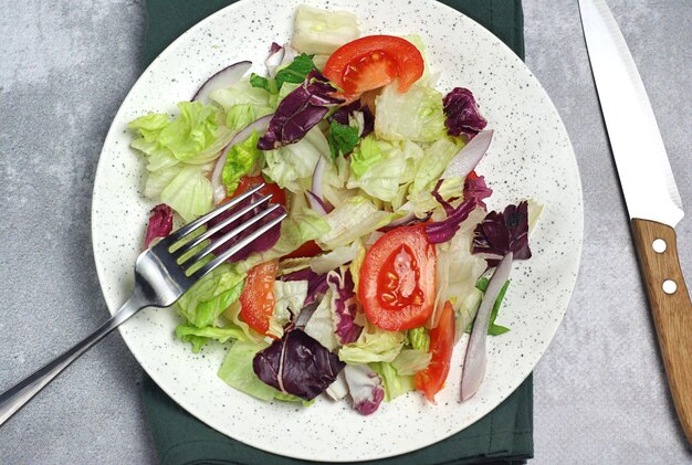 Salad with tomatoes leaves in a plate on the table There is a fork and a knife nearby There is a towel under the plate