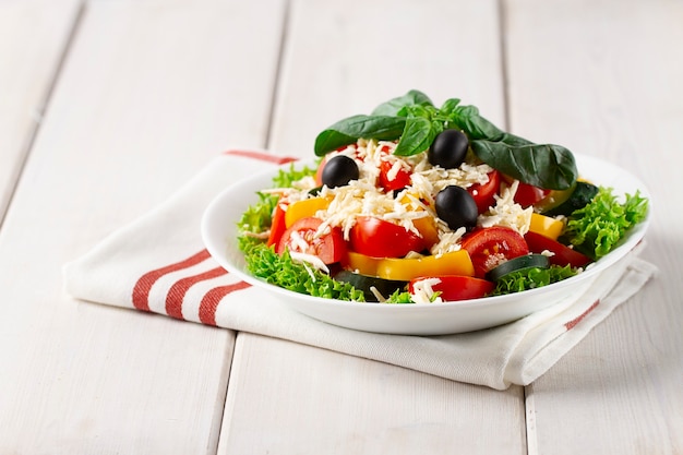 Salad with vegetables and cheese, on a white plate over a kitchen cloth on white wooden background