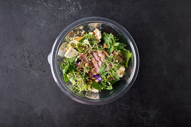 Salade met gerookte eendenborstgroenten, kaas en kruiden in een transparante container op dark on