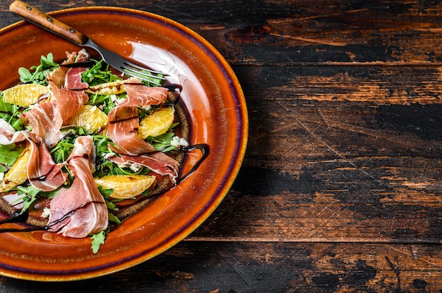 Salade met jamonham, parmezaanse kaas, rucola en mandarijn op een bord. Donkere houten tafel. Bovenaanzicht.