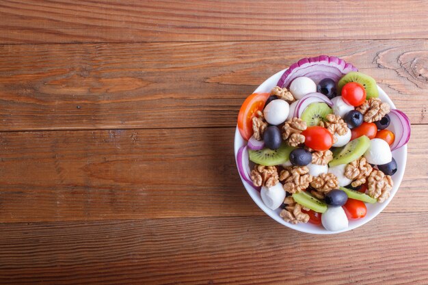 Salade met kersentomaten, mozarellakaas, olijven, kiwi, en okkernoten op bruine houten achtergrond.