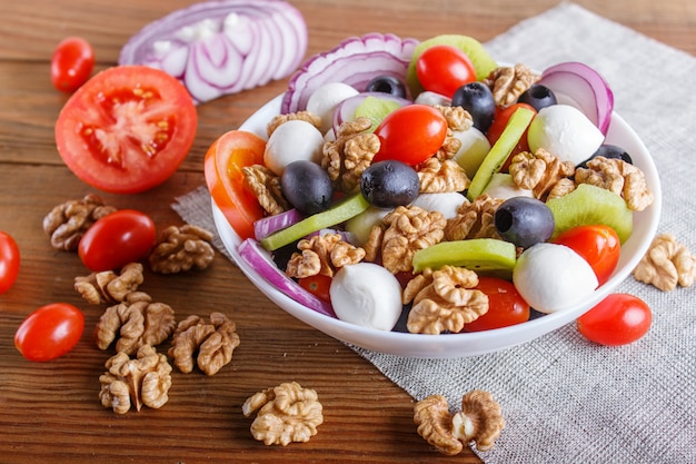 Salade met kersentomaten, mozarellakaas, olijven, kiwi, en okkernoten op bruine houten achtergrond.