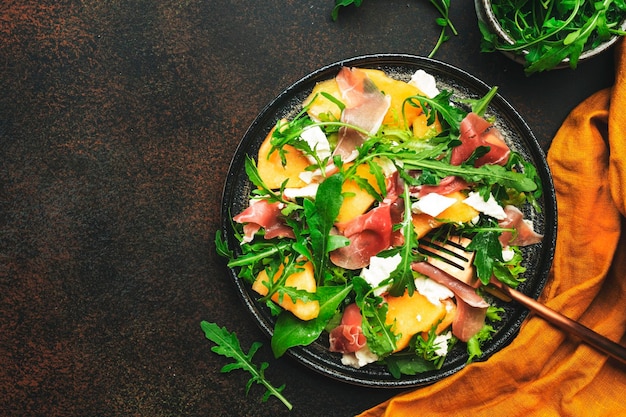 Salade met meloen jamon geitenkaas en rucola op bruine achtergrond bovenaanzicht kopie ruimte