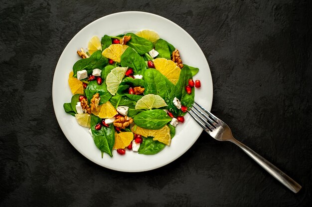 salade met spinazie, sinaasappel, citroen, granaatappel, fetokaas, walnoot op een witte plaat tegen de achtergrond van leisteen, steen of beton