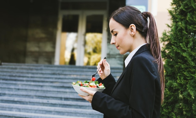 Salade op zakenlunch Portret van een mooie moderne en glimlachende zakenvrouw in een zwart pak met een bord groentesalade in de handen die ze op straat eet in de buurt van het werk