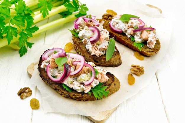 Salade van zalm, bladsteel selderij, rozijnen, walnoten, rode uien en kwark op geroosterd brood met groene sla op papier op een lichte houten plank achtergrond