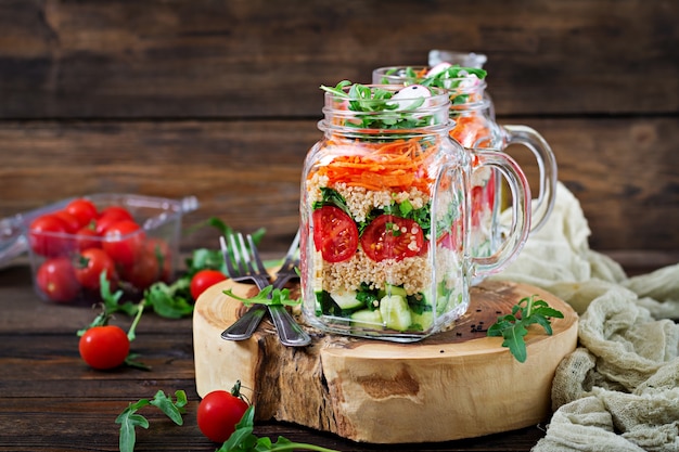 Insalate con quinoa, rucola, ravanello, pomodori e cetriolo in barattoli di vetro sulla tavola di legno. cibo sano, dieta, disintossicazione e concetto vegetariano